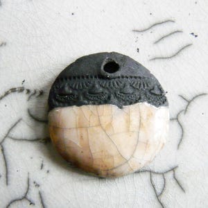 Può includere: Un ciondolo in ceramica fatto a mano con un design a semicerchio. La metà superiore è grigio scuro con un motivo strutturato, e la metà inferiore è bianco sporco con un aspetto screpolato e invecchiato. Ha un foro per l'infilatura.