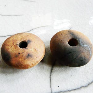 Peut inclure: Deux perles rondes en forme de donut, percées d'un trou central. Les perles présentent un mélange de tons bruns et beiges, avec des accents brun foncé. Elles sont probablement en pierre ou en argile, pour la création de bijoux.