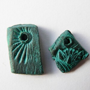 Może przedstawiać: Dwa małe, prostokątne, zielone metalowe zawieszki z otworami w środku. Jedna zawieszka ma okrągły wzór z promieniującymi liniami, druga ma bardziej abstrakcyjny wzór.