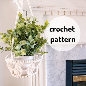 May include: A white crocheted hanging planter with a green plant inside. The planter is hanging from a white ceiling against a white wall with a fireplace. The text "crochet pattern" is written in a white circle.