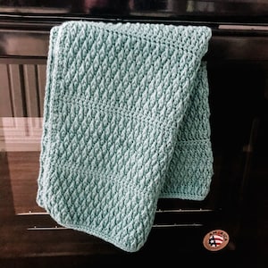 May include: A light teal crocheted kitchen towel with a textured pattern, hanging on a black oven door. The towel is folded over, showcasing the detailed stitch work. A small "America" sticker is visible.