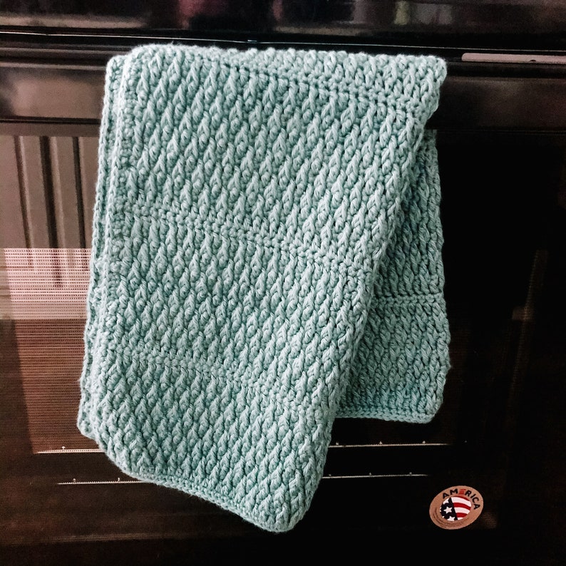 May include: A light teal crocheted kitchen towel with a textured pattern, hanging on a black oven door. The towel is folded over, showcasing the detailed stitch work. A small "America" sticker is visible.