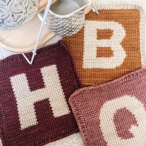 May include: Three crocheted squares with the letters H, B, and O in white yarn on a burgundy, mustard yellow, and pink background.