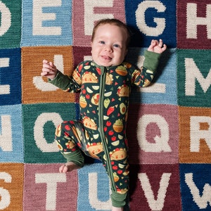 May include: A baby wearing a green and black patterned onesie with a zipper is lying on a colorful alphabet play mat. The baby is smiling and looking at the camera.