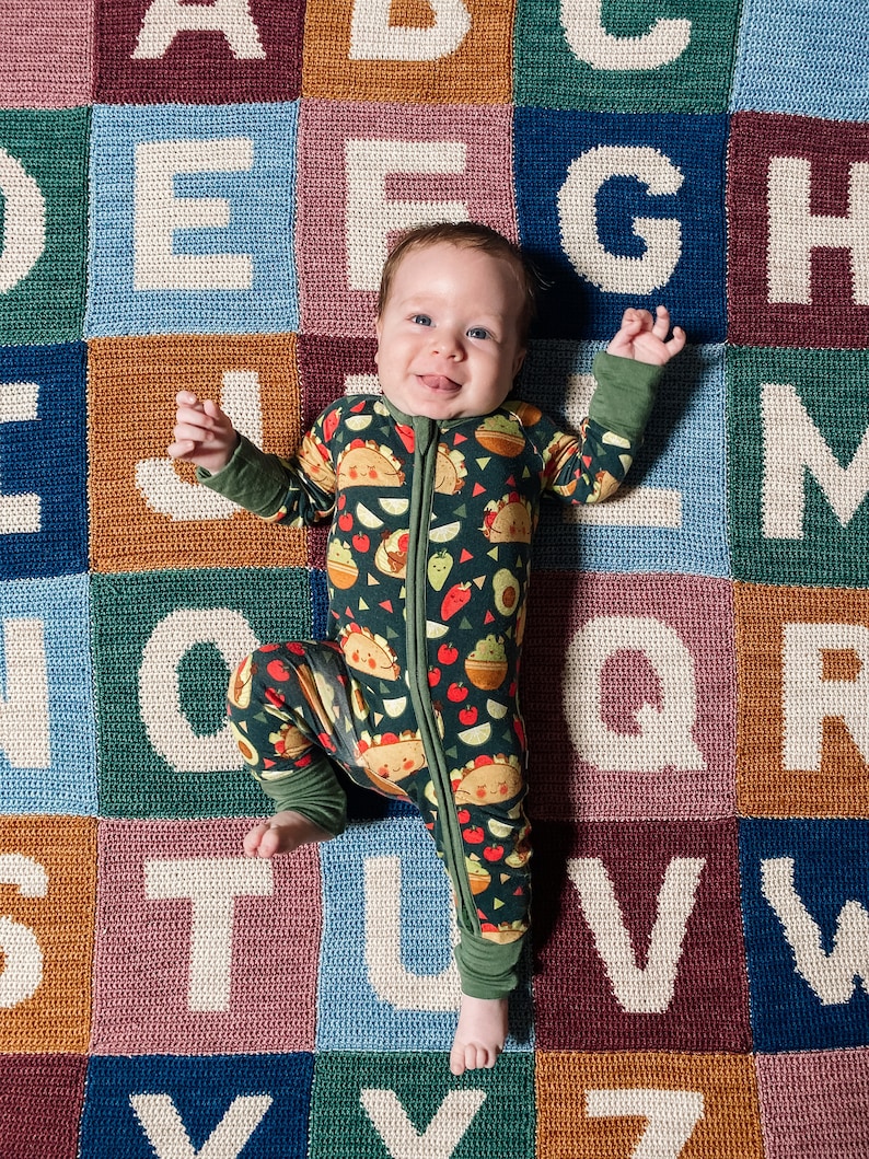 May include: A baby wearing a green and black patterned onesie with a zipper is lying on a colorful alphabet play mat. The baby is smiling and looking at the camera.