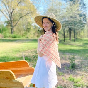 May include: A woman wearing a white dress and a pink and white crocheted shawl stands in a grassy field. She is wearing a wide-brimmed straw hat and looking over her shoulder at the camera.