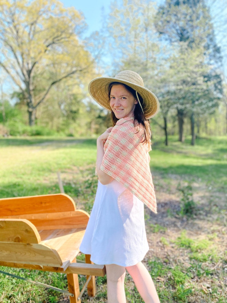 May include: A woman wearing a white dress and a pink and white crocheted shawl stands in a grassy field. She is wearing a wide-brimmed straw hat and looking over her shoulder at the camera.