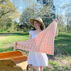 May include: A woman wearing a white dress and a straw hat holds a pink and white crocheted shawl in a grassy field.