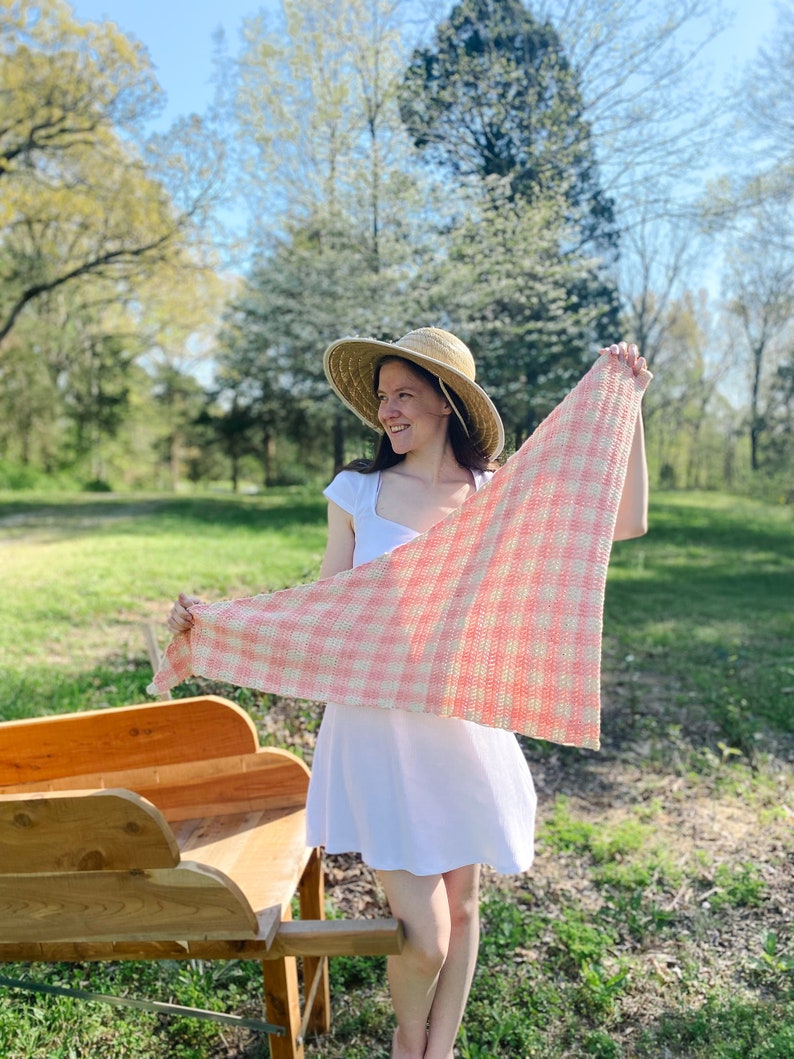 May include: A woman wearing a white dress and a straw hat holds a pink and white crocheted shawl in a grassy field.