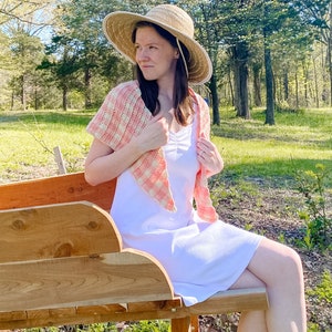 May include: A woman wearing a white dress and a pink and white gingham crochet shawl sits on a wooden bench outdoors. She is wearing a straw sun hat and looking off to the side.