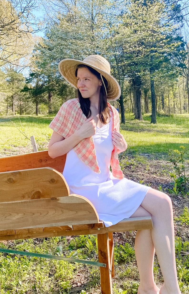 May include: A woman wearing a white dress and a pink and white gingham crochet shawl sits on a wooden bench outdoors. She is wearing a straw sun hat and looking off to the side.