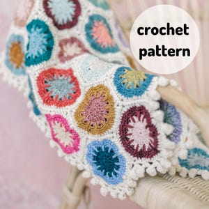 May include: A colorful crochet blanket with a white border and a crocheted flower pattern. The blanket is draped over a wicker chair.