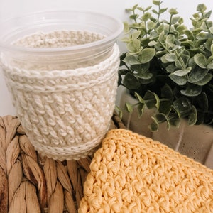 May include: A close-up of two crocheted cup cozies. One is a cream-colored cozy with a simple stitch pattern. The other is a yellow cozy with a more intricate stitch pattern. Both cozies are made with soft yarn and are perfect for keeping your hands warm while enjoying your favorite beverage.