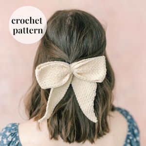 May include: A crocheted bow hair accessory in a cream color. The bow is tied in a large, loose knot and has long, flowing tails.