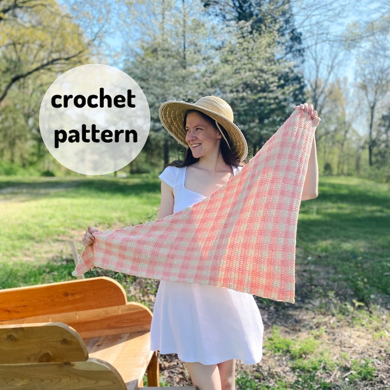 May include: A woman wearing a white dress and a straw hat holds up a pink and white crochet shawl. The shawl has a gingham pattern. The text "crochet pattern" is written in a circle in the upper left corner of the image.