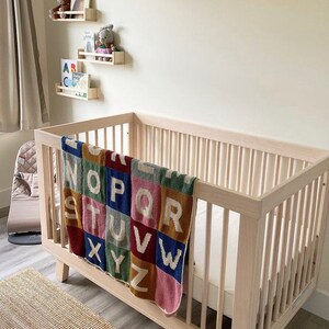 May include: A white wooden crib with a colorful alphabet blanket draped over the side. The blanket features a patchwork design with each letter in a different color and pattern.
