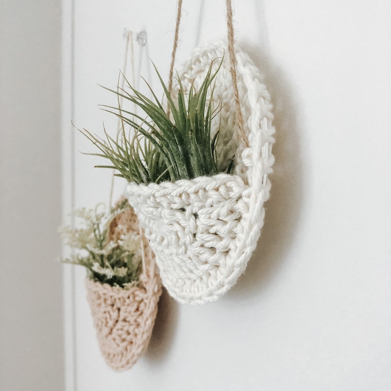 May include: Two white crocheted hanging plant pots with green air plants inside. The pots are hanging from a white wall.