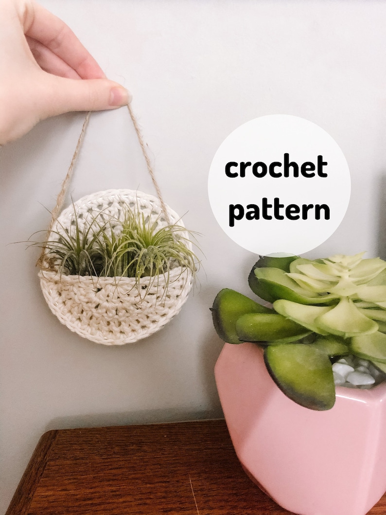 May include: A white crocheted hanging planter with two air plants inside. The planter is hanging from a twine cord and is attached to a white wall. The text "crochet pattern" is written in black on a white circle.