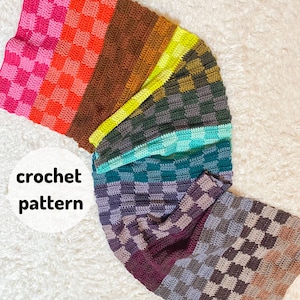 May include: A colourful crochet blanket with a checkered pattern in shades of pink, brown, green, and blue. The blanket is folded in a crescent shape, with the different coloured stripes visible.