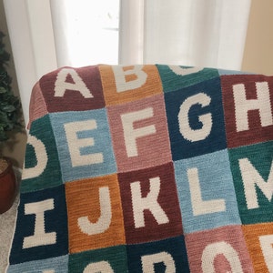 May include: A colorful crocheted blanket with the letters of the alphabet in white. Each letter is in a different colored square.