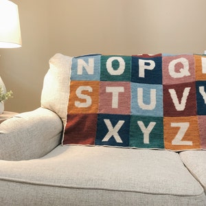 May include: A colorful crocheted alphabet blanket with the letters N through Z in white on a couch.