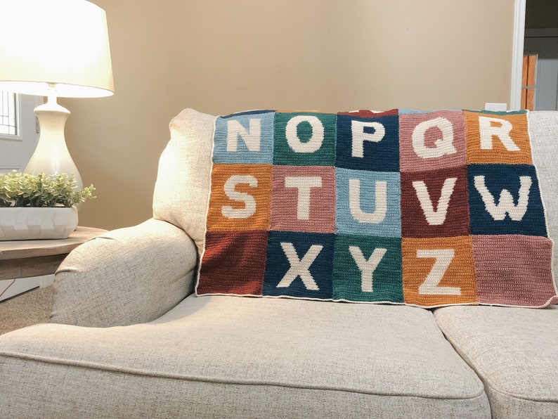 May include: A colorful crocheted alphabet blanket with the letters N through Z in white on a couch.