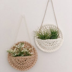 May include: Two macrame plant hangers with a white and a brown crocheted pot. The white pot has two green air plants inside. The brown pot has a small bouquet of white flowers inside.