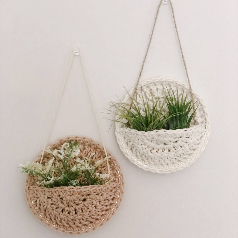 May include: Two macrame plant hangers with a white and a brown crocheted pot. The white pot has two green air plants inside. The brown pot has a small bouquet of white flowers inside.