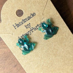 May include: A pair of translucent dark green frog-shaped stud earrings attached to a light brown cardboard display card. The card features the printed text "Handmade by ArtseyFartsy" in black ink, with a circular hole at the top for hanging.