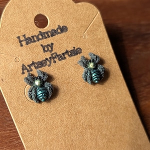 May include: A pair of spider-shaped stud earrings featuring a textured dark gray body, a metallic teal abdomen, and a small iridescent green bead at the center. The earrings are attached to a brown cardboard display card printed with the text Handmade by ArtseyFartsie.