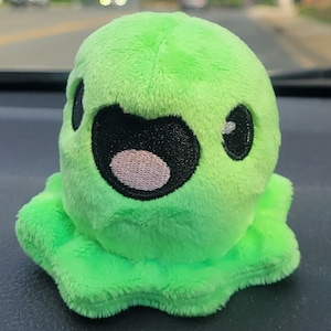May include: A plush green creature with a wide open mouth and large black eyes sits on a car dashboard. The plush toy has a ruffled base and a light pink mouth. The background shows a road and trees.