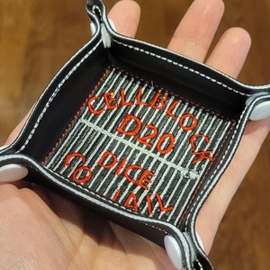 May include: Black leather dice tray with white stitching and white snaps. The tray has a black and white striped fabric lining with red embroidered text that reads "Cell Block D20 Dice Tray".