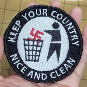 Op de afbeelding: Ronde patch met de tekst "KEEP YOUR COUNTRY NICE AND CLEAN" rond een witte cirkel. Het midden toont een persoon die een rood hakenkruis in een vuilnisbak gooit. De patch heeft een zwarte rand.