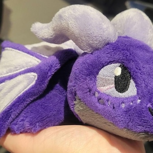 Puede incluir: Un peluche de dragón en tonos morados, grises y blancos. El dragón tiene rasgos faciales bordados, incluyendo un ojo grande y pequeños detalles. Las alas son blancas con ribetes morados. Un juguete suave y tierno.