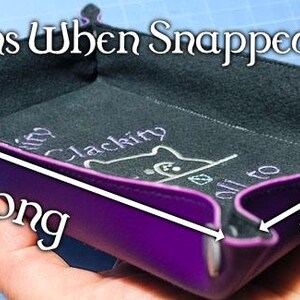 K&ouml;nnte beinhalten: Ein lila W&uuml;rfel-Tablett mit schwarzem Innenraum, das im zusammengeklappten Zustand 17,78 cm lang und 12,7 cm breit ist. Das Tablett tr&auml;gt den Text "Dimensions When Snapped Into Tray" und "Clackity" mit einer Katzen-Grafik.