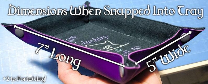 K&ouml;nnte beinhalten: Ein lila W&uuml;rfel-Tablett mit schwarzem Innenraum, das im zusammengeklappten Zustand 17,78 cm lang und 12,7 cm breit ist. Das Tablett tr&auml;gt den Text "Dimensions When Snapped Into Tray" und "Clackity" mit einer Katzen-Grafik.