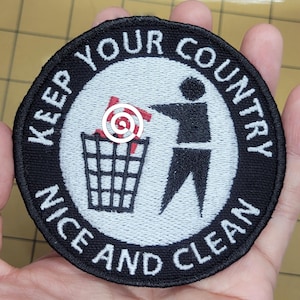May include: A circular embroidered patch with a black border and white background. It features a black silhouette of a person placing a red swastika into a black trash can. The text "KEEP YOUR COUNTRY" and "NICE AND CLEAN" is embroidered in white.