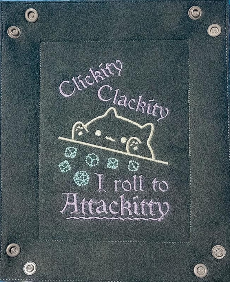 K&ouml;nnte beinhalten: Ein dunkelblaugr&uuml;nes W&uuml;rfelspieltablett mit einem gestickten Design. Das Design zeigt eine Katze und die Worte "Clickity Clackity" und "I roll to AttacKitty" in einem hellen Lila. Das Tablett hat Metallschnappverschl&uuml;sse.