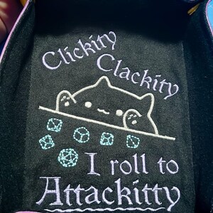 K&ouml;nnte beinhalten: Schwarzes W&uuml;rfelspieltablett mit lila Einfassung und dem Text "Clickity Clackity" und "I roll to Attackitty" in Lavendel. Eine wei&szlig;e Katzenzeichnung und hellblaue W&uuml;rfel sind ebenfalls abgebildet.