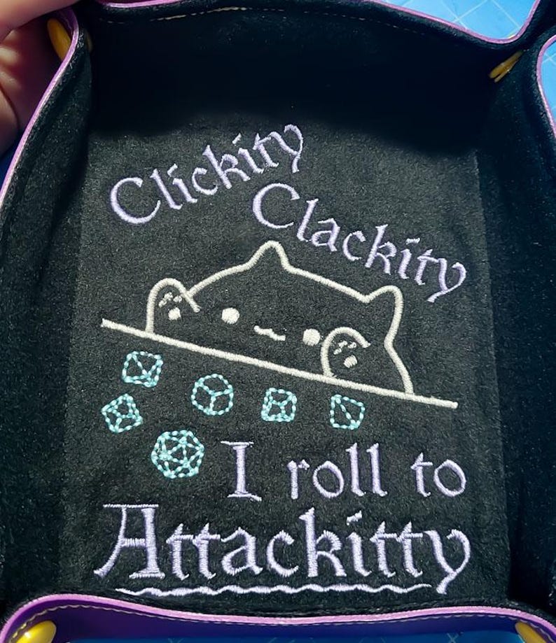 K&ouml;nnte beinhalten: Schwarzes W&uuml;rfelspieltablett mit lila Einfassung und dem Text "Clickity Clackity" und "I roll to Attackitty" in Lavendel. Eine wei&szlig;e Katzenzeichnung und hellblaue W&uuml;rfel sind ebenfalls abgebildet.
