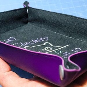 K&ouml;nnte beinhalten: Eine rautenf&ouml;rmige, violette Ablage mit schwarzem Innenraum. Die Ablage hat einen gen&auml;hten, violetten Rand und wird mit grauen Druckkn&ouml;pfen zusammengehalten. Der Innenraum zeigt wei&szlig;en, gestickten Text und eine Katzenzeichnung. Die Ablage ist f&uuml;r kleine Gegenst&auml;nde konzipiert.