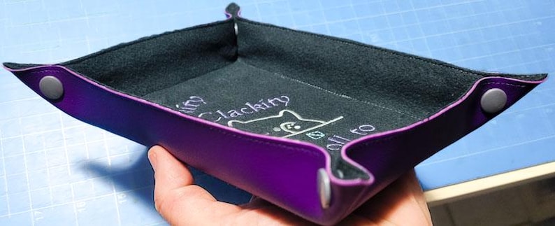 K&ouml;nnte beinhalten: Eine rautenf&ouml;rmige, violette Ablage mit schwarzem Innenraum. Die Ablage hat einen gen&auml;hten, violetten Rand und wird mit grauen Druckkn&ouml;pfen zusammengehalten. Der Innenraum zeigt wei&szlig;en, gestickten Text und eine Katzenzeichnung. Die Ablage ist f&uuml;r kleine Gegenst&auml;nde konzipiert.