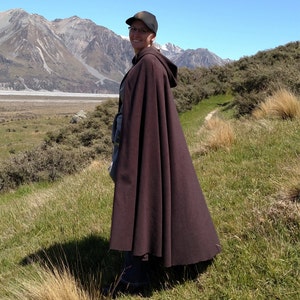 May include: A person wearing a brown hooded cloak stands in a grassy field with mountains in the background.