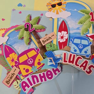 May include: A colorful beach-themed cake topper with a sun wearing sunglasses, surfboards, a blue VW bus, and palm trees. The topper includes the name "LUCAS" and the number "2". The words "way to beach" are on a wooden sign.