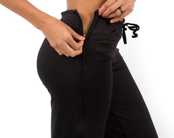 May include: Black athletic pants with a wide leg design. The pants feature a zipper detail on the side and a drawstring waistband. The model is wearing gray sneakers. The pants are made of a smooth, stretchy material.