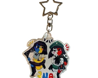 Könnte beinhalten: Ein klarer Acryl-Schlüsselanhänger mit einer Cartoon-Illustration von zwei Figuren und dem Wort "ENA" in bunten Buchstaben. Der Schlüsselanhänger hat einen sternförmigen Metallverschluss und eine silberne Kette.