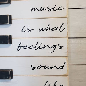 Puede incluir: Un letrero de madera blanco con detalles de teclas de piano negras. El letrero muestra la cita "la m&uacute;sica es lo que los sentimientos suenan" en escritura cursiva negra. Un art&iacute;culo decorativo.