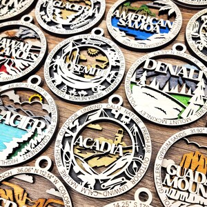 Puede incluir: Una colecci&oacute;n de adornos de madera que representan varios parques nacionales y monumentos. Cada adorno est&aacute; pintado con colores vibrantes e incluye el nombre del parque o monumento, junto con sus coordenadas geogr&aacute;ficas. Los adornos est&aacute;n dispuestos sobre una superficie de madera.