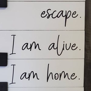 Puede incluir: Letrero de madera blanco con texto negro: "escape. I am alive. I am home." El letrero est&aacute; hecho de tablas horizontales y tiene herrajes negros. El texto est&aacute; en una fuente cursiva.
