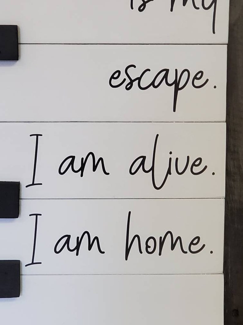 Puede incluir: Letrero de madera blanco con texto negro: "escape. I am alive. I am home." El letrero est&aacute; hecho de tablas horizontales y tiene herrajes negros. El texto est&aacute; en una fuente cursiva.